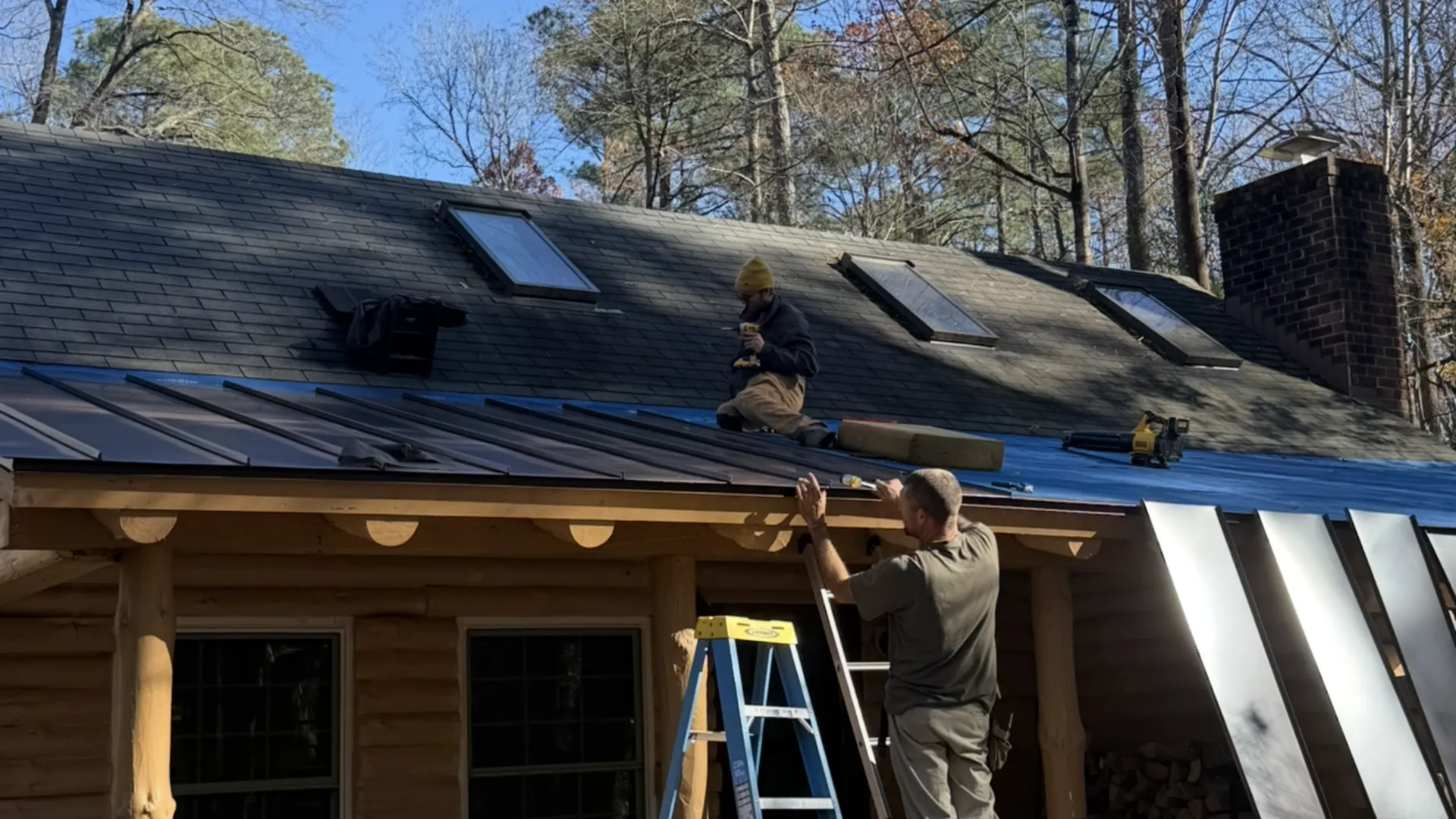 Two men installing a new metal roof over blue underlayment and shingles on a log cabin house.